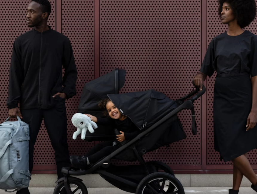 Adult in dark clothing leaning over a black stroller to tend to a child reclined in the seat against a deep red wall, a stroller for growing families.