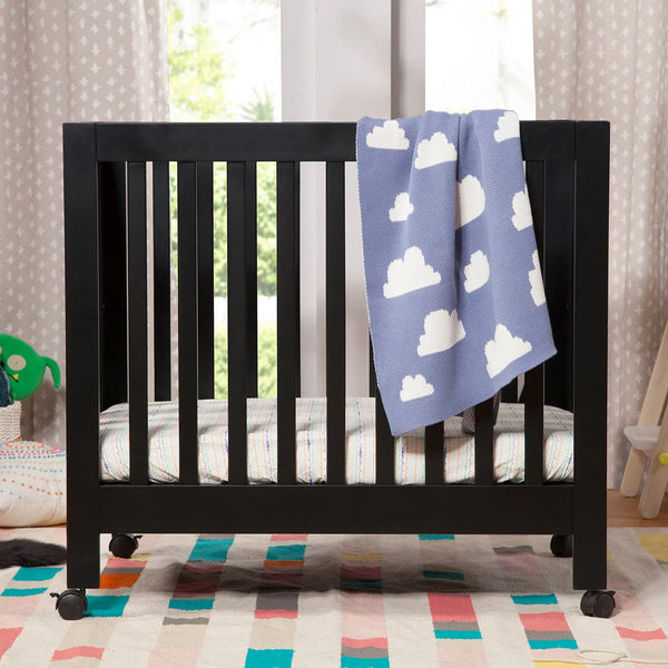 Babyletto Origami mini crib in black on caster wheels with a cloud blanket in a bright nursery. 