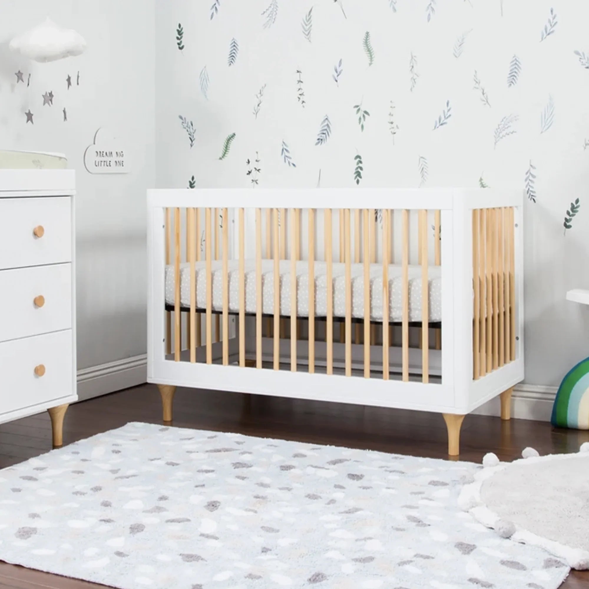 Convertible crib with a white frame and natural wood spindles in a botanical wallpaper nursery, paired with a matching white three-drawer dresser. 