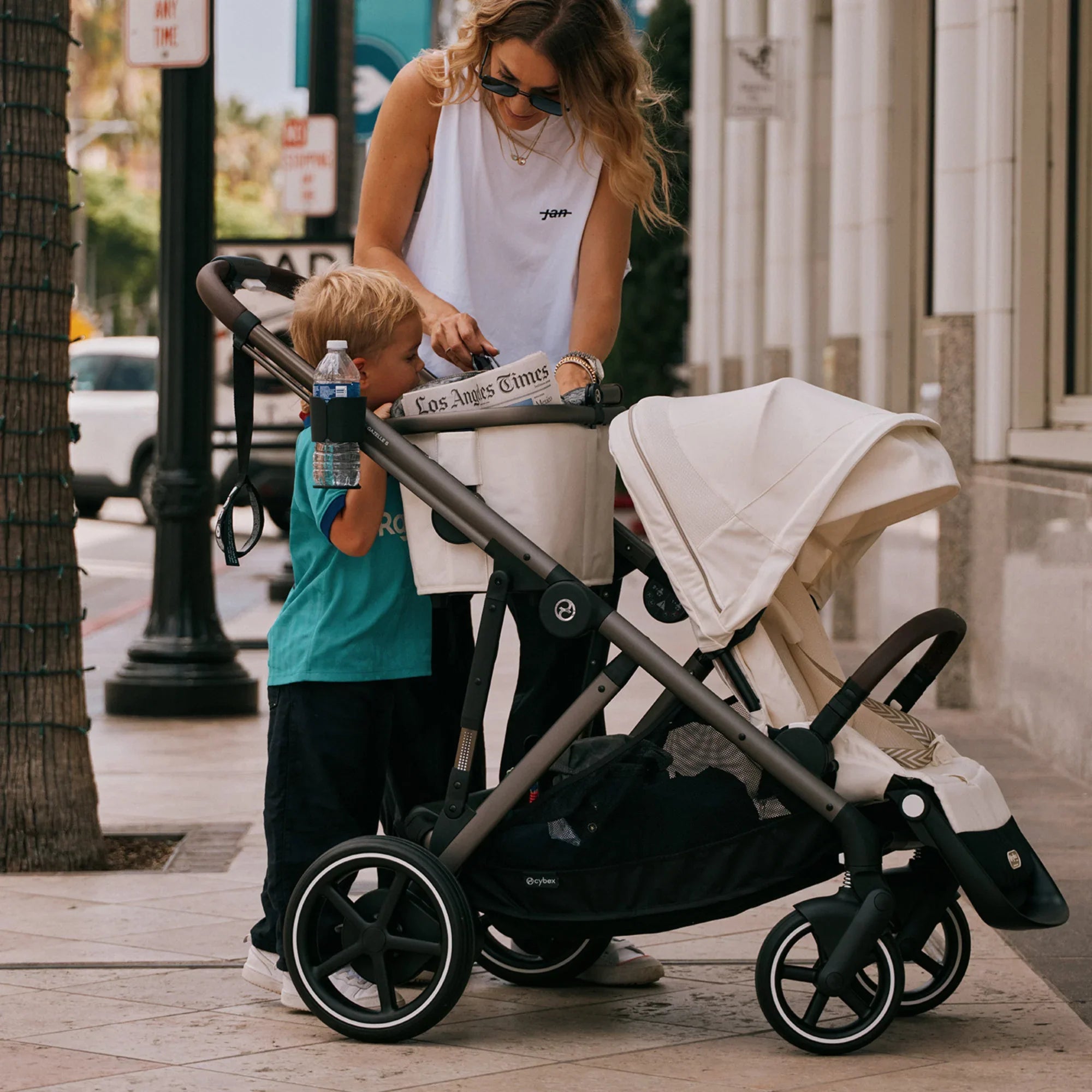 A woman pauses on a city sidewalk to organize items in a stroller while a young child stands beside her, highlighting everyday urban parenting moments