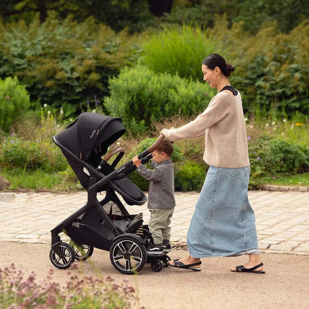 Mother pushes Nuna DEMI Next stroller with toddler rider board in a park 