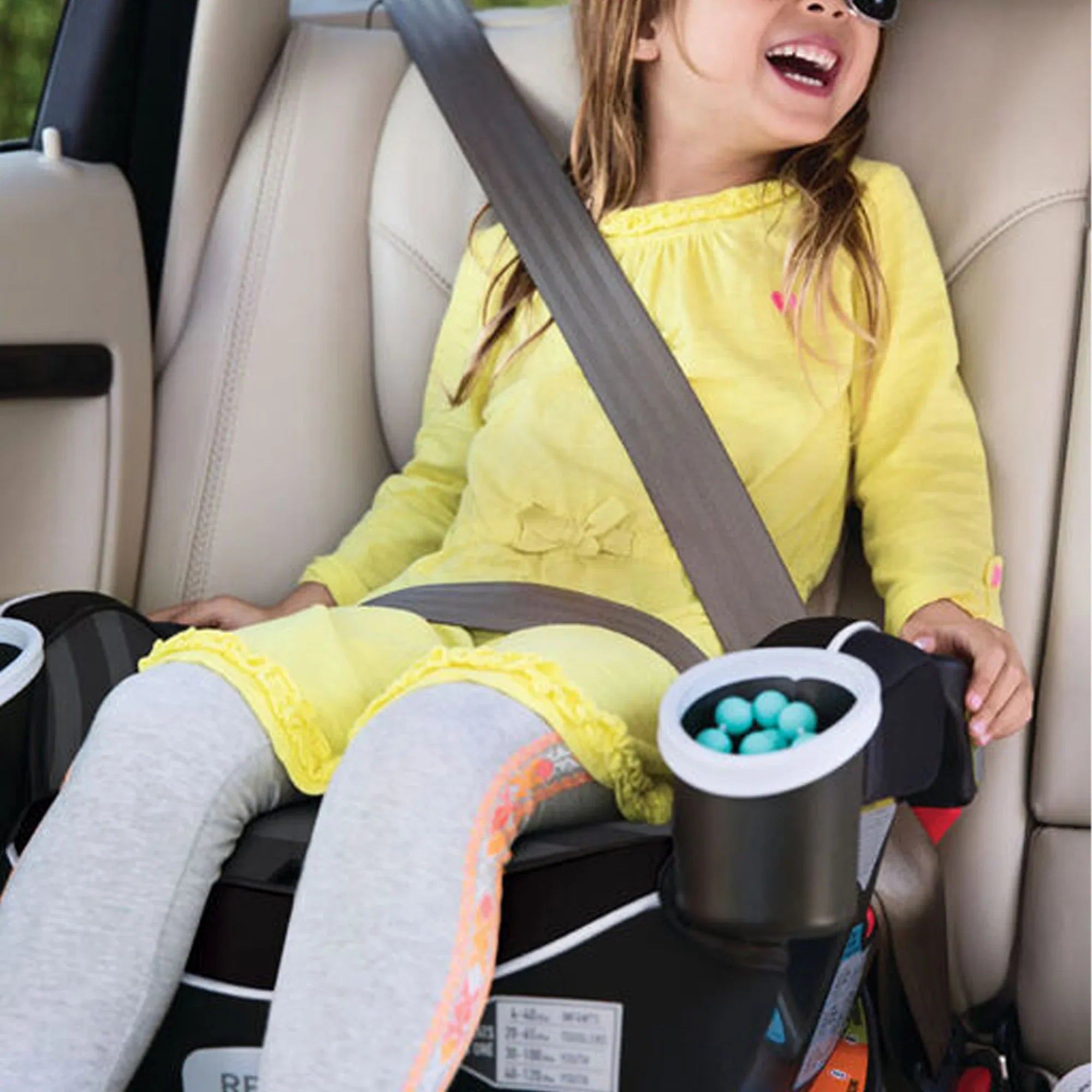 A laughing child in sunglasses rides in a high-back booster seat, secured by the vehicle's diagonal seat belt, with a snack cup at hand.