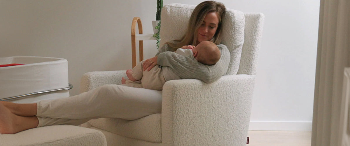 Mom cuddling her baby in white glider chair with ottoman in serene, minimalist nursery.