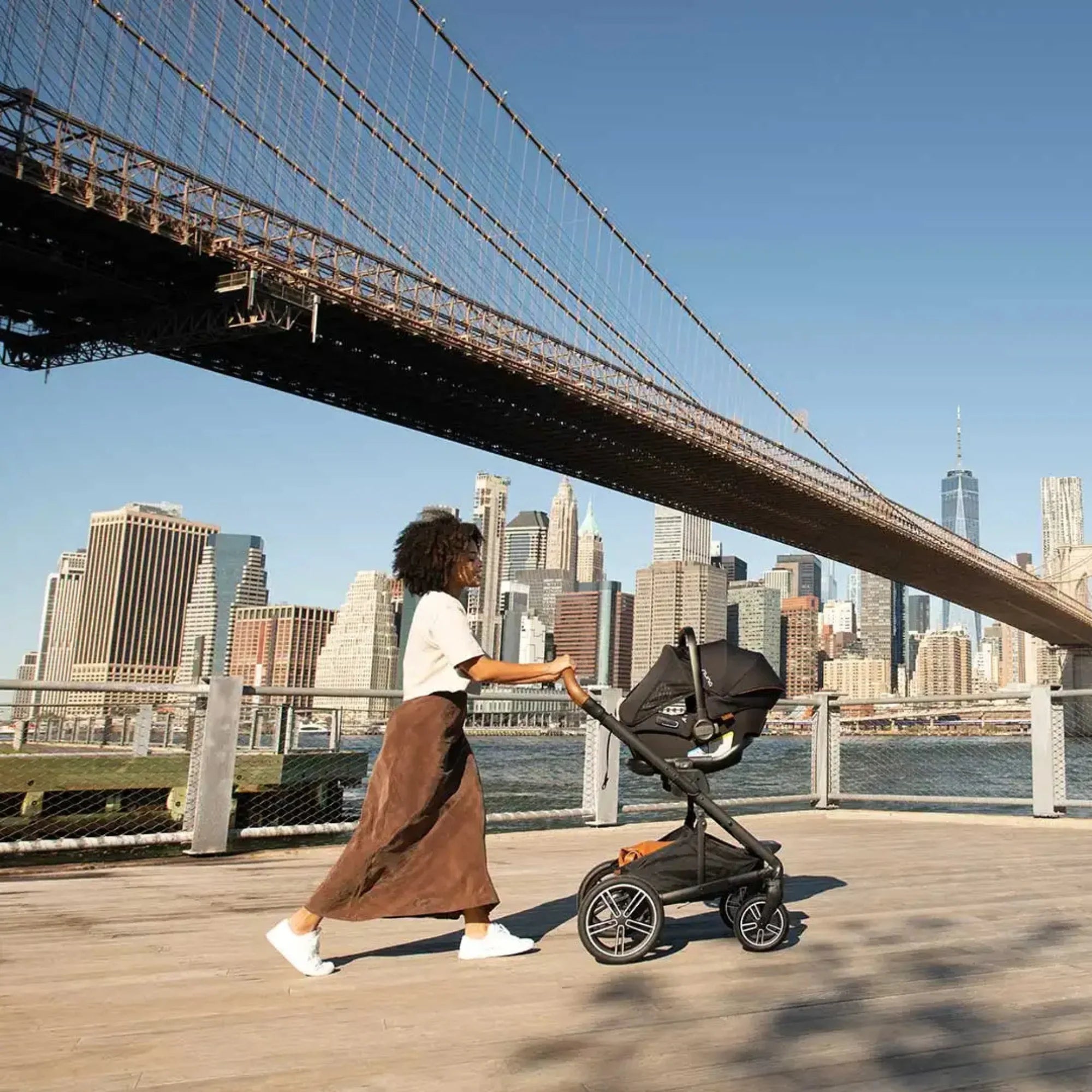 Parent pushing a stroller with a car seat combo along a waterfront path, with a city skyline and suspension bridge in the background.