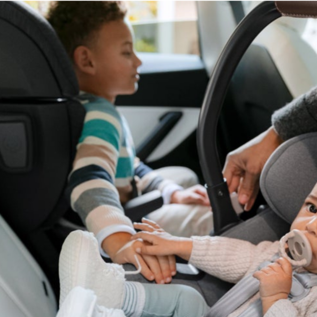 A Toddler and a baby holding hands in the backseat while seated in car seats.
