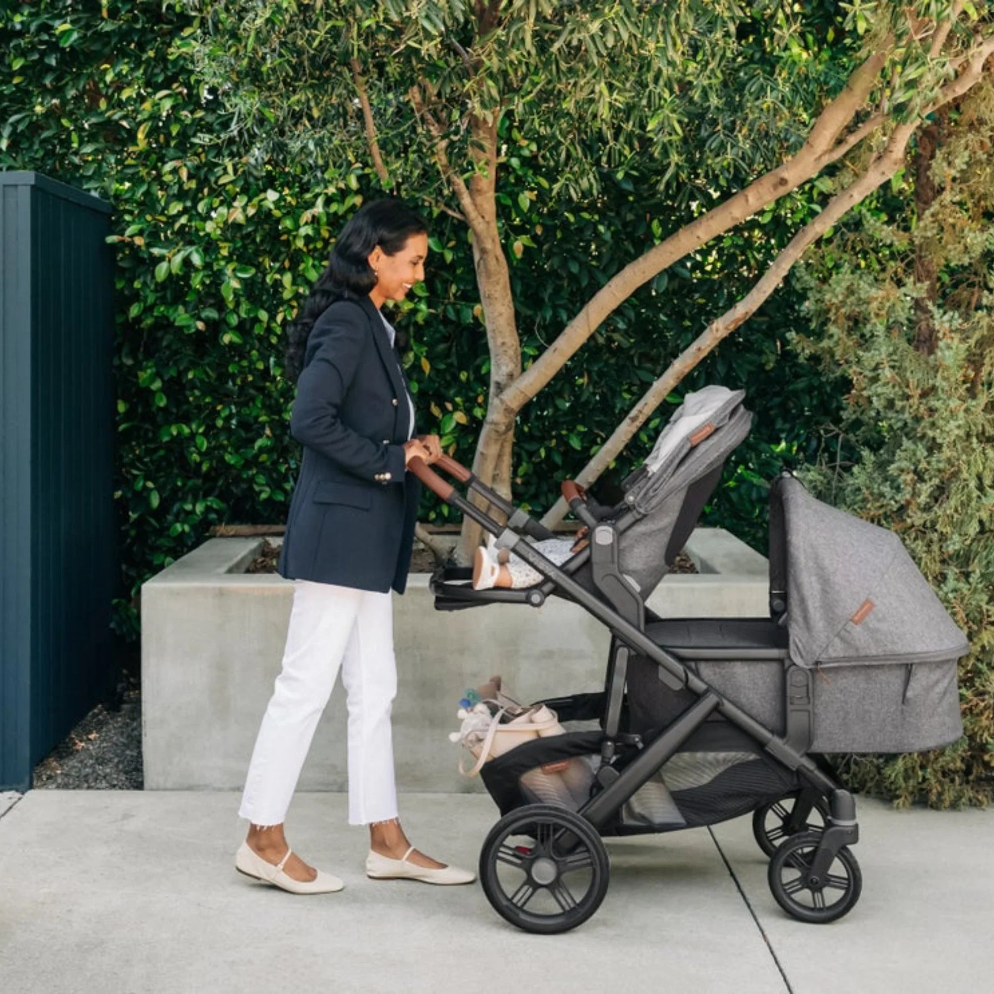  A parent in a navy blazer pushes a grey UPPAbaby Vista set up as a double, a toddler seat plus a second seat/bassinet on the black frame with tan leather handle, outdoors by a hedge.