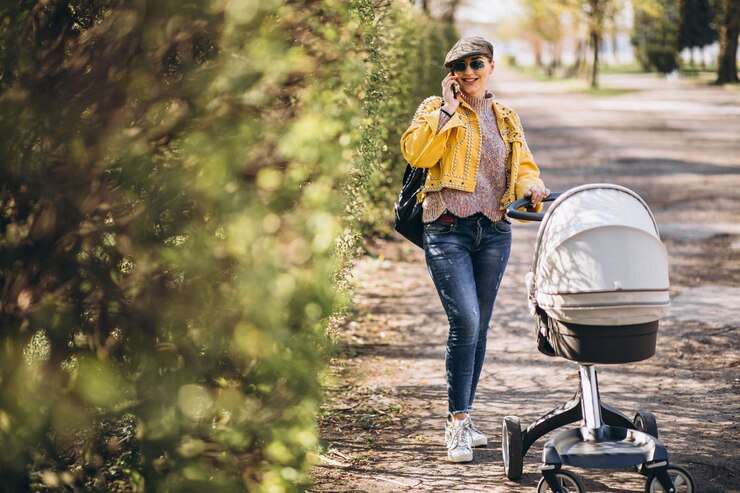 Smiling woman pushing a newborn stroller on a sunny day while talking on the phone in a park, wearing a yellow jacket and casual outfit.