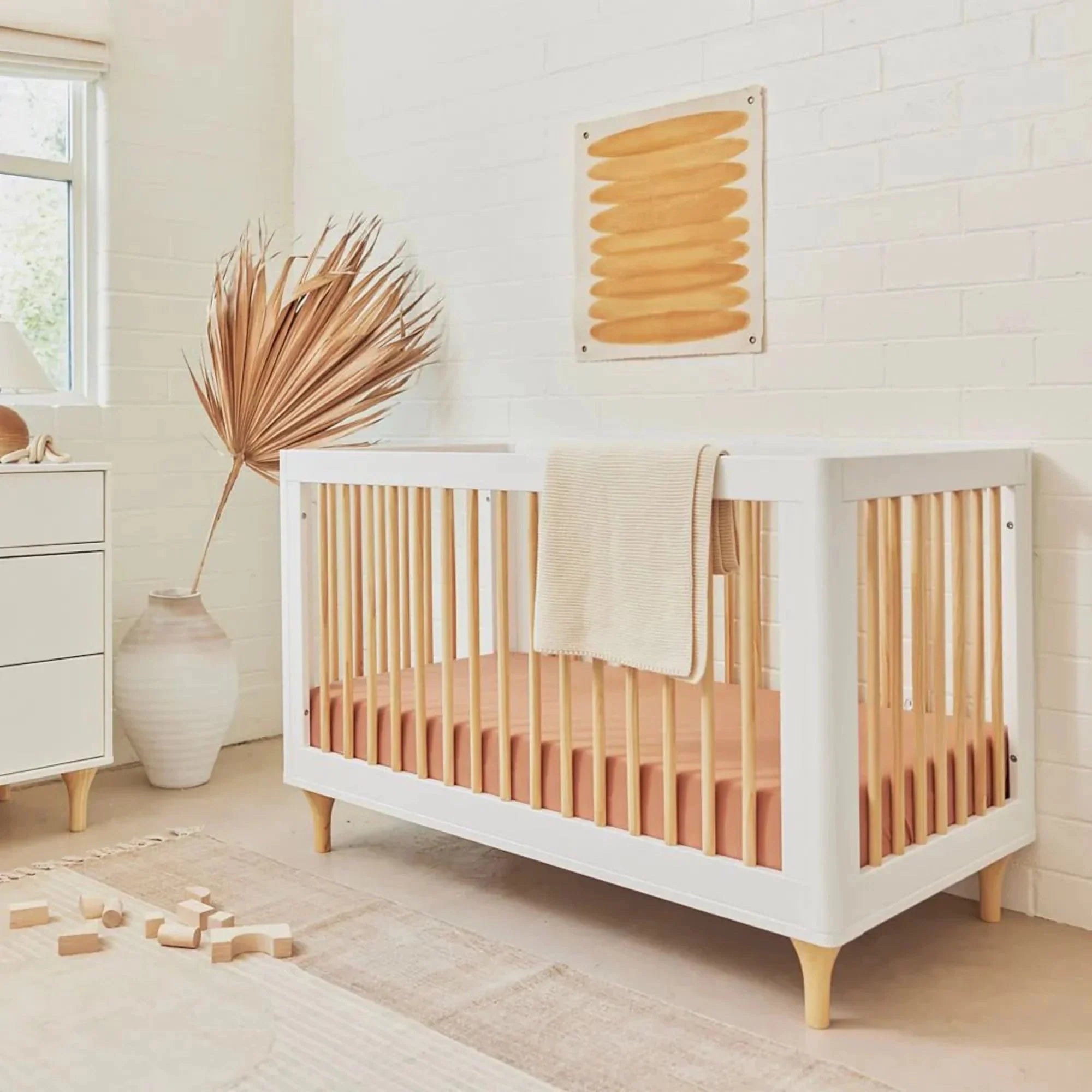 Mid-century modern baby crib with white frame and natural wood spindles, styled in a bright neutral nursery with a terracotta fitted sheet. 