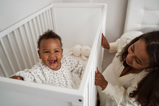 A joyful baby laughing in a white crib with a mother smiling beside them.