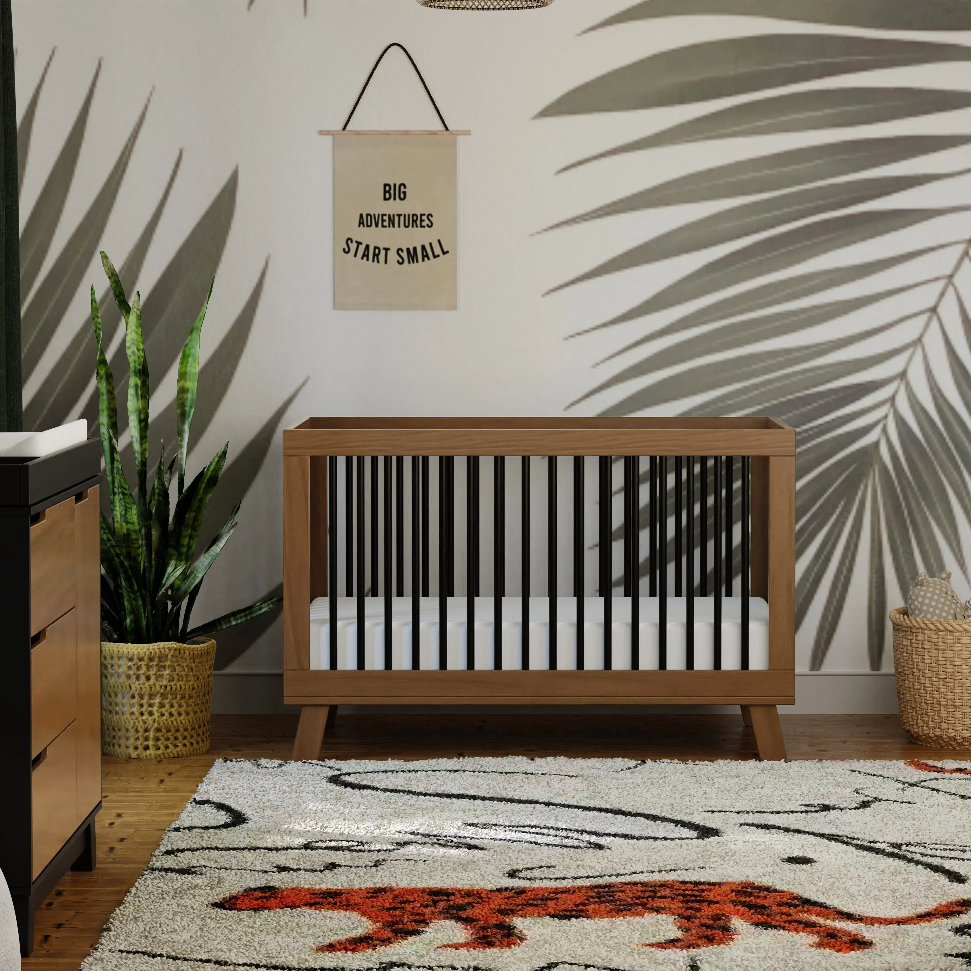  Babyletto Hudson crib in a two-tone finish against a palm-leaf mural nursery, with a dresser, snake plant, woven basket, "Big Adventures Start Small" wall banner, and a tiger-motif rug.
