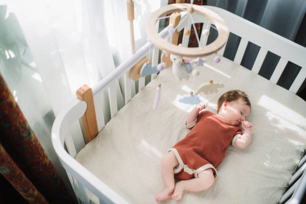 A newborn baby sleeping on their back in a white slatted crib with a wooden and felt crib mobile.