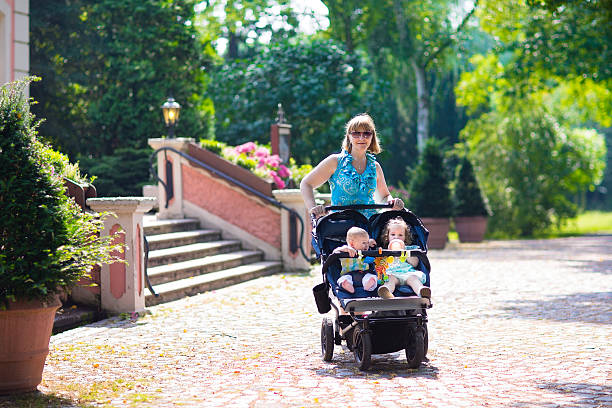 A woman pushing a double stroller with two young children through a park on a sunny day.