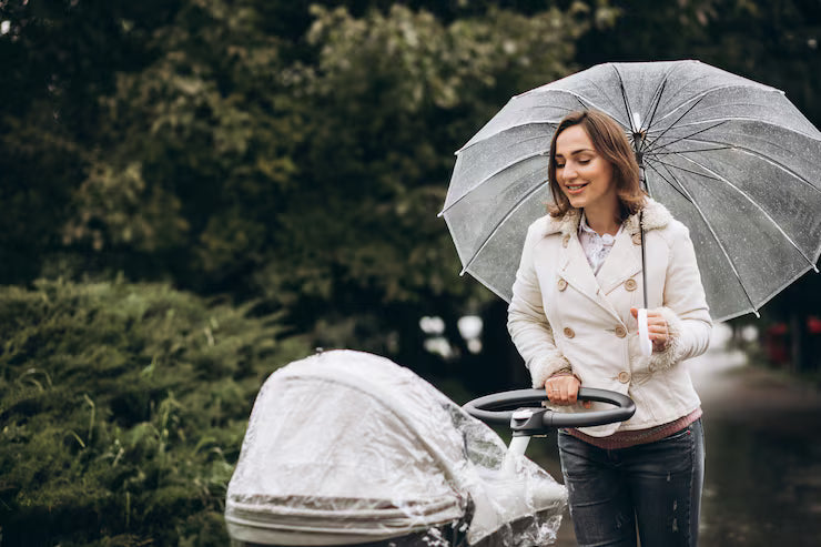 A mother walking in a rainy park with a baby stroller covered by a rain cover, holding a transparent umbrella.