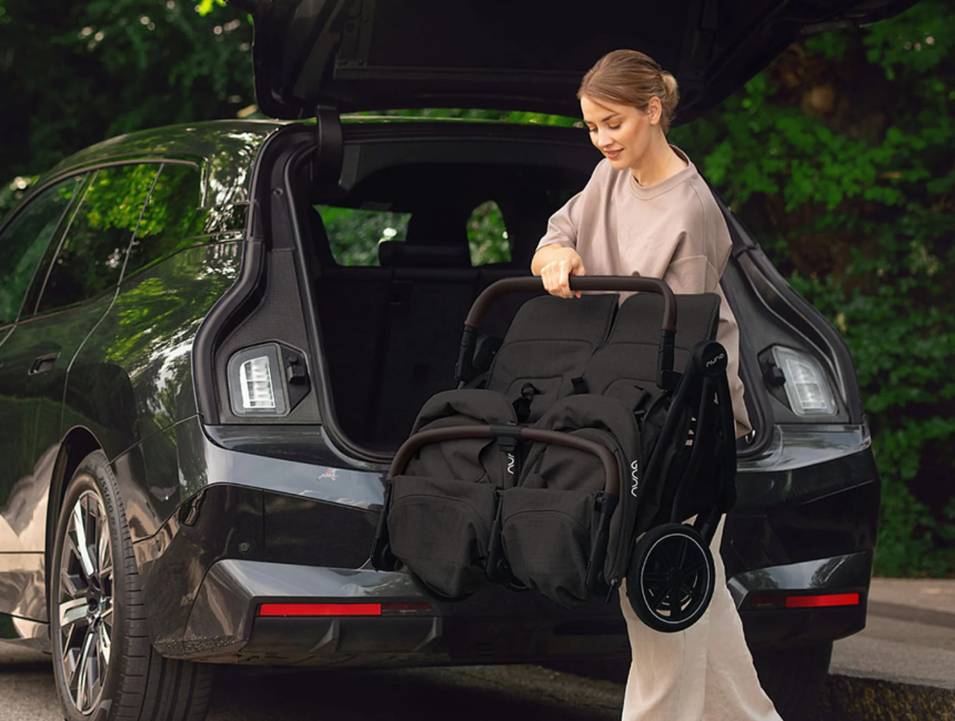 Parent standing at the open trunk of a dark car, holding a folded black stroller, a car seat compatible stroller for family trips by car.