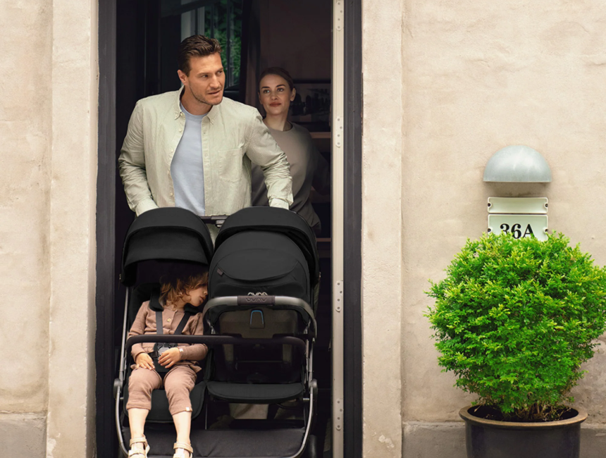 Adult standing in a doorway beside a black double stroller with two young children seated inside, next to a potted shrub outside.