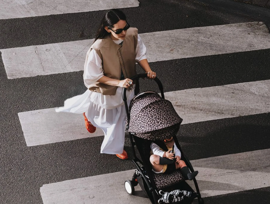 Parent in a white dress and tan vest crosses a striped crosswalk pushing a Stokke YOYO³ stroller with the Newborn Pack attached