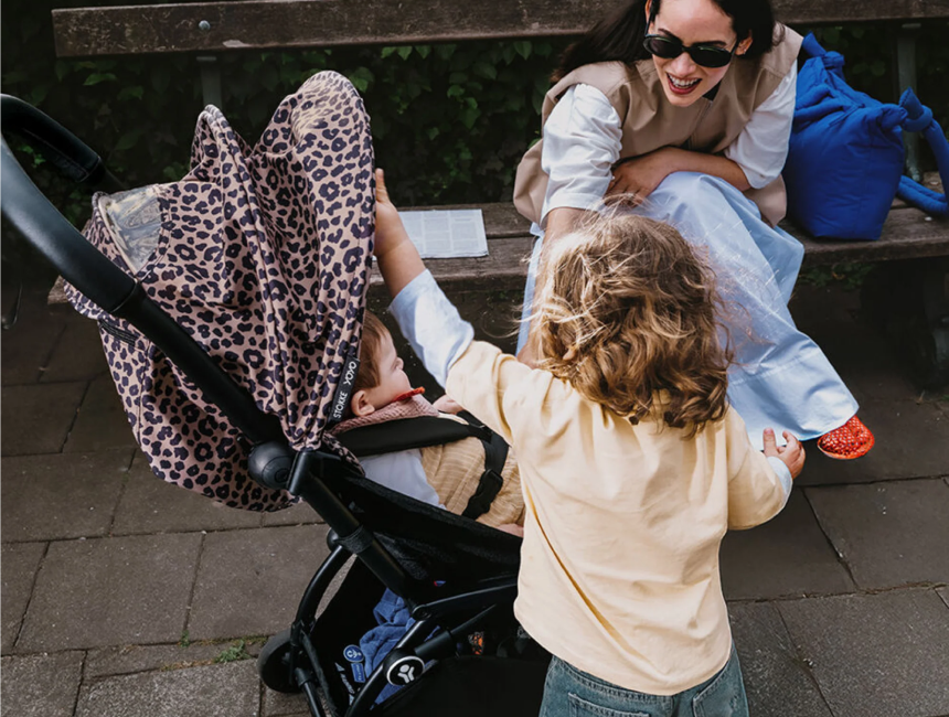 Parent wearing sunglasses sits on a park bench holding an infant while a toddler reaches over, stroller with floral canopy nearby
