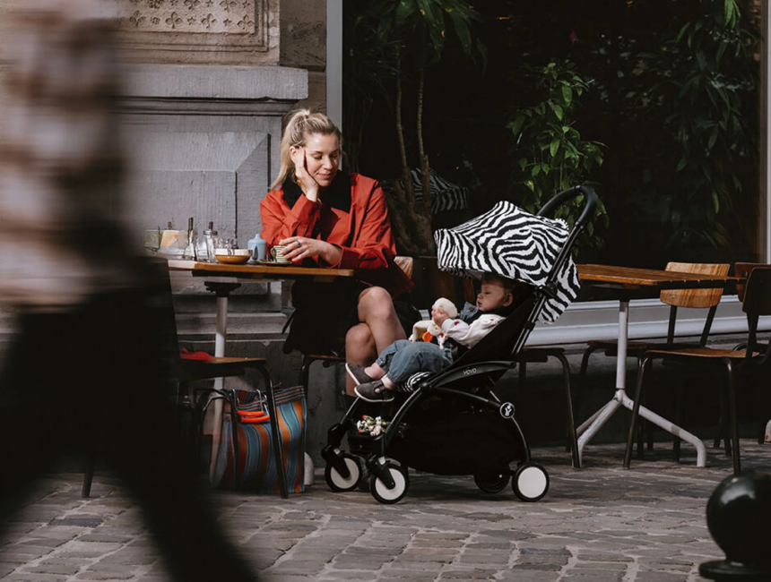 Parent in an orange jacket seated at an outdoor cafe table beside a Stokke YOYO³ stroller with a checkered Newborn Pack canopy