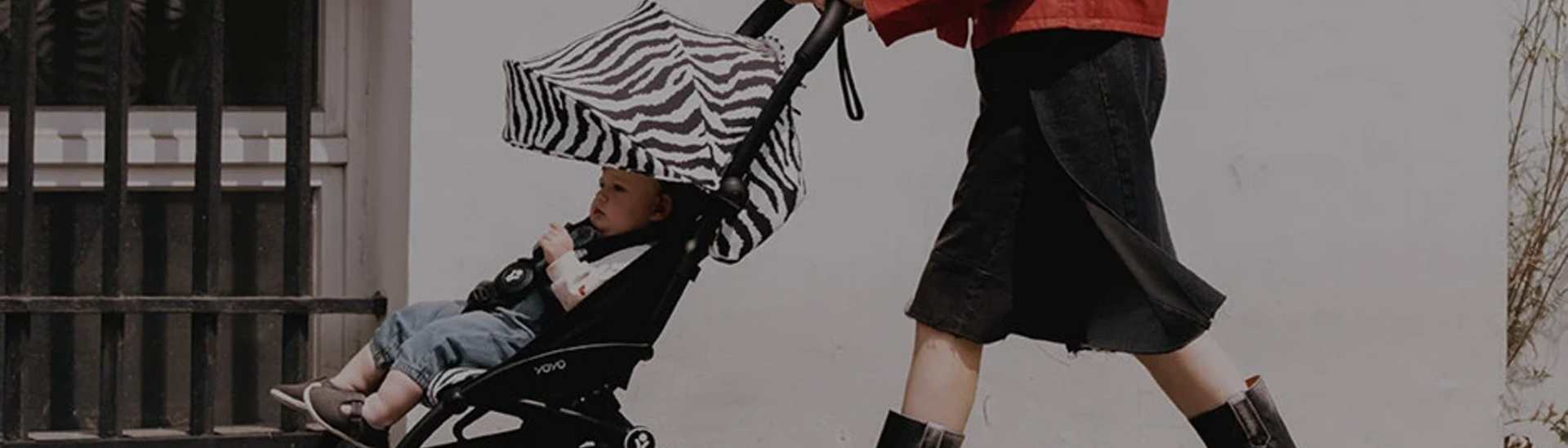A parent walks along a city sidewalk pushing a Stokke YOYO³ stroller fitted with a bold zebra-print Newborn Pack canopy cover