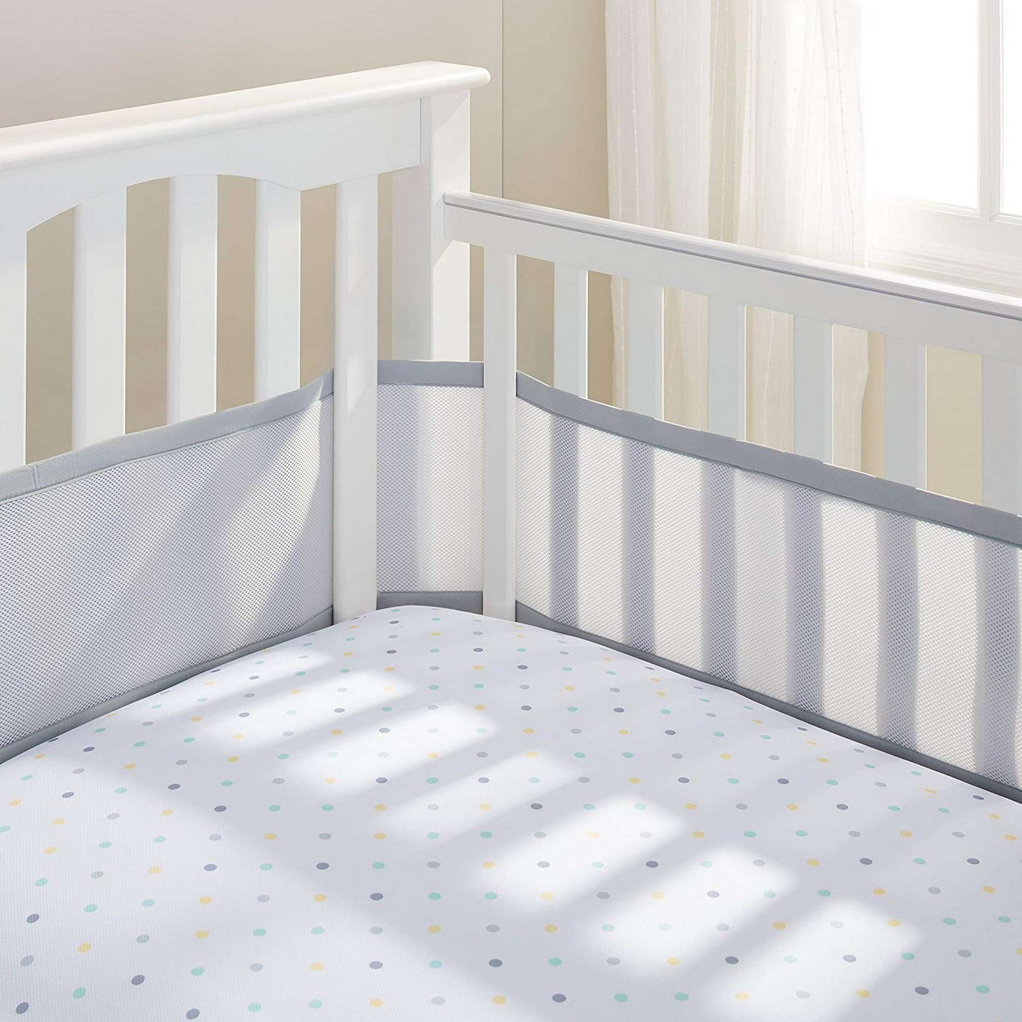 White baby crib with soft grey mesh crib liner and polka-dot fitted sheet, placed by a sunlit window for a calm nursery setting.